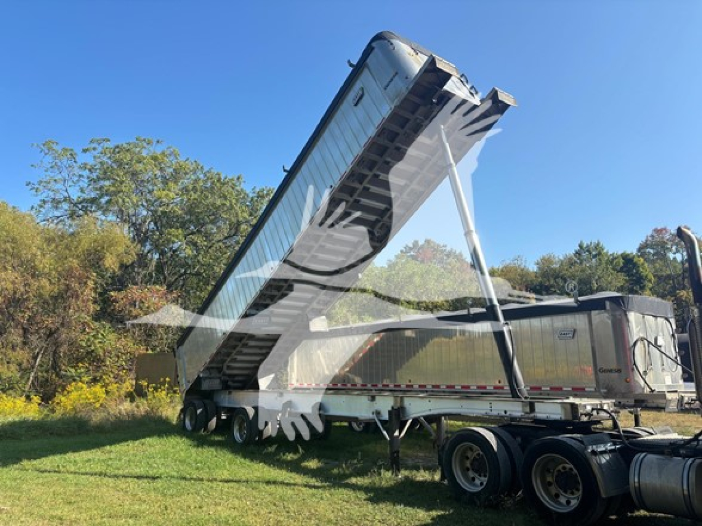 Trailer hitch and hydraulic hoist of a dump trailer raised in the air, graffiti on side, blue sky, trees in background.