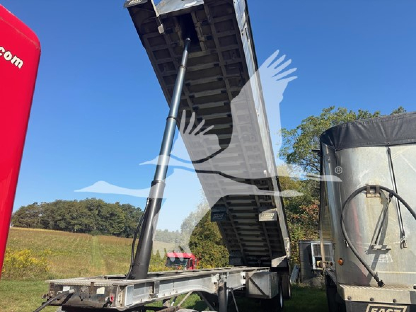 Trailer/Dump trailer in raised position showing hydraulic piston and frame components. Visible text on structures is minimal or not legible in the image.