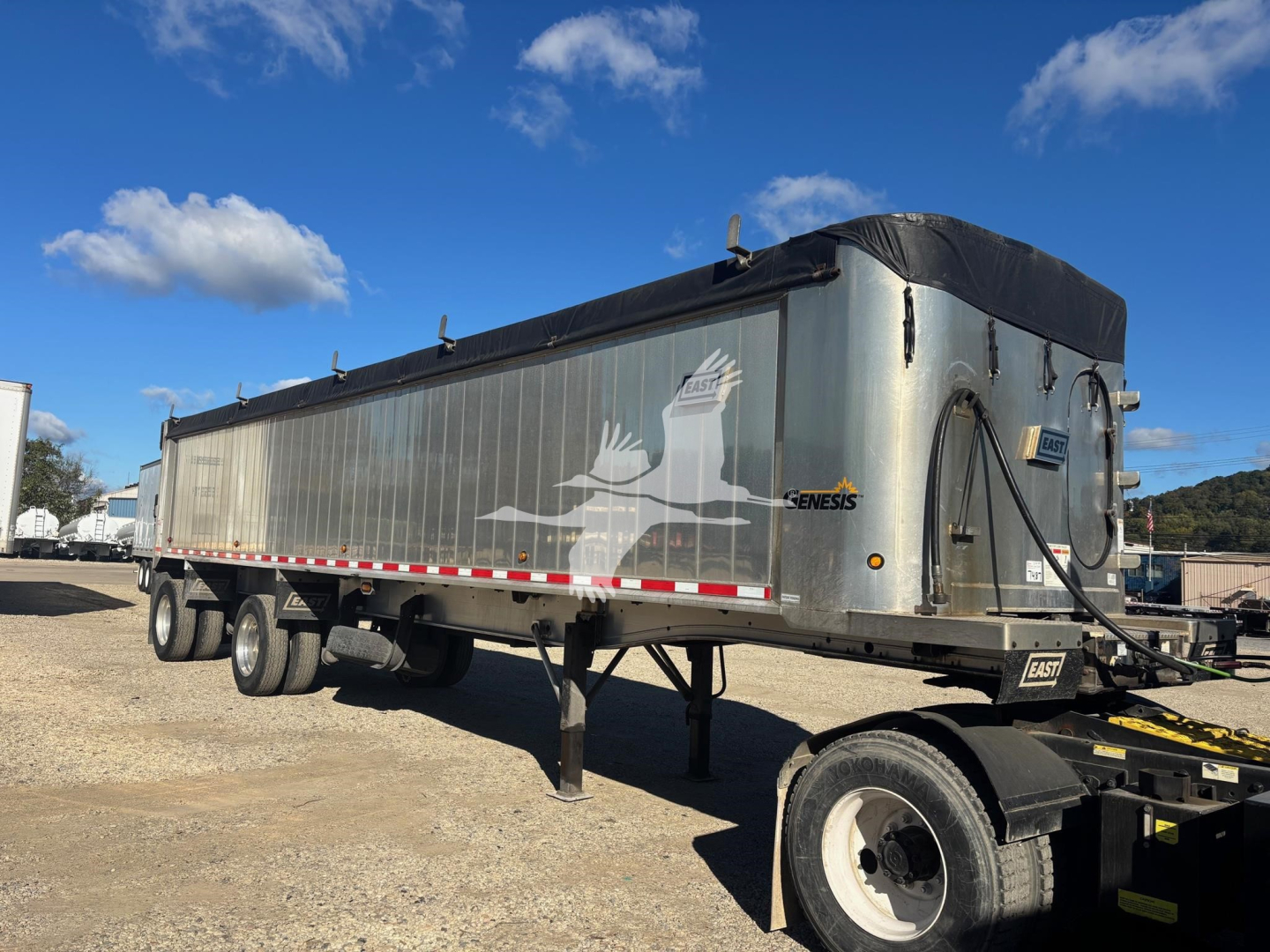 Trailer showing Empire East Hybrid dump trailer with silver sides, black tarp, Genesis logo, and dual rear axles; side view with clear blue sky.