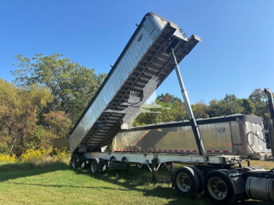 Trailer dump bed raised on hydraulic strut with semi-trailer in background, dual axles, metal frame, black and silver colors.