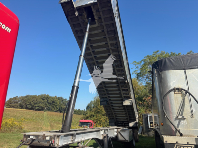 Dump trailer with hydraulic dump bed raised, hydraulic strut visible, exposed undercarriage, road field background.