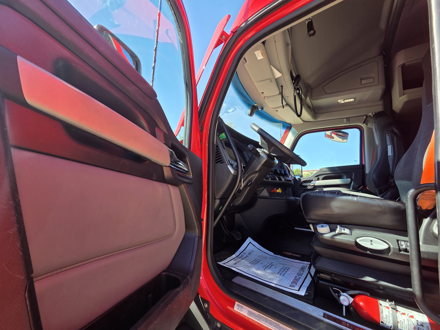 Truck interior door open showing red exterior with tan/cream door panel and driver's seat; dashboard and steering wheel visible; paper documents on floor; overhead controls and center console.