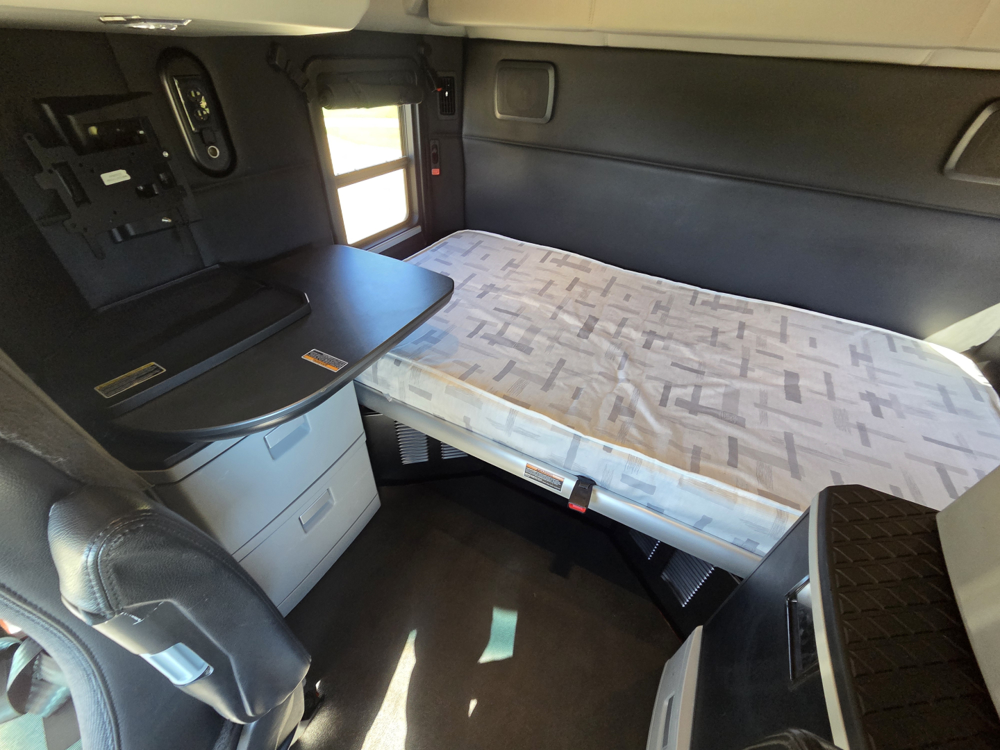 Kenworth T680 sleeper cab interior with folded bed and overhead compartments; front passenger seat area visible; small desk surface with drawers and control panel.
