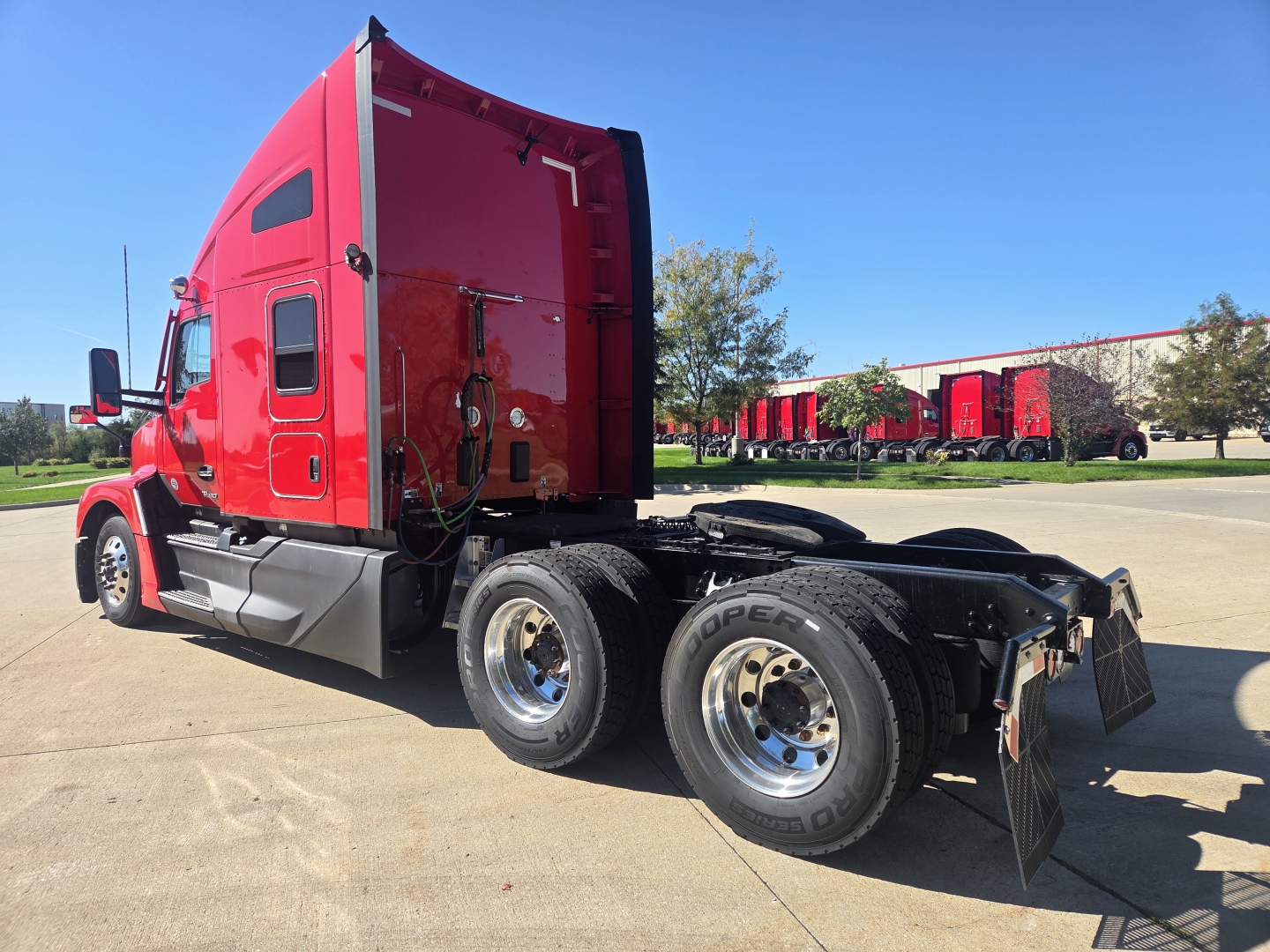 Kenworth T680 tractor, red cab, side view showing dual rear wheels and fifth wheel, Linked fuel lines and exhaust visible on a concrete lot.