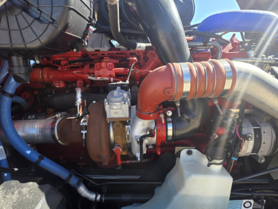 Engine bay of a 2023 Kenworth T680 with orange turbocharger pipe, red engine components, blue hoses, and white coolant reservoir cap visible.