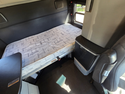 Kenworth T680 sleeper cab interior with single bed and storage compartments, light fabric mattress, door window, and dashboard area visible.