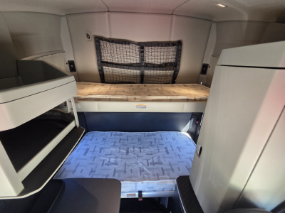 Truck sleeper cab interior showing bunk area with storage shelves and a mesh netted window. Stocked with light wood workspace and blue patterned floor mat.