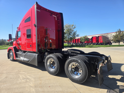 Kenworth T680 tractor, red cab, side view showing dual rear wheels and fifth wheel, Linked fuel lines and exhaust visible on a concrete lot.