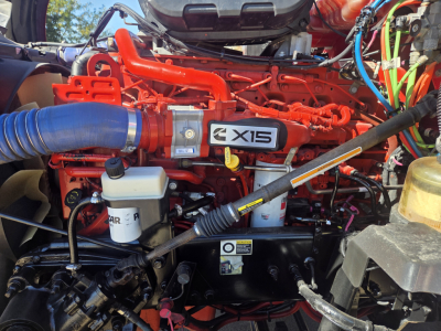 Kenworth T680 engine bay showing X15 badge, red engine components, blue intake hose, coolant reservoir, and various hoses and wires.