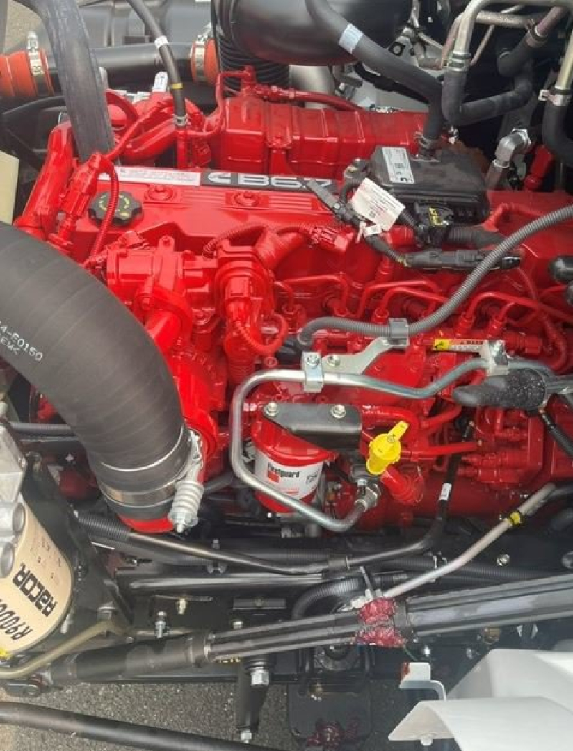 Engine bay of a new Hino L6 cab chassis showing a bright red engine with hoses, fittings, and a yellow dipstick label.