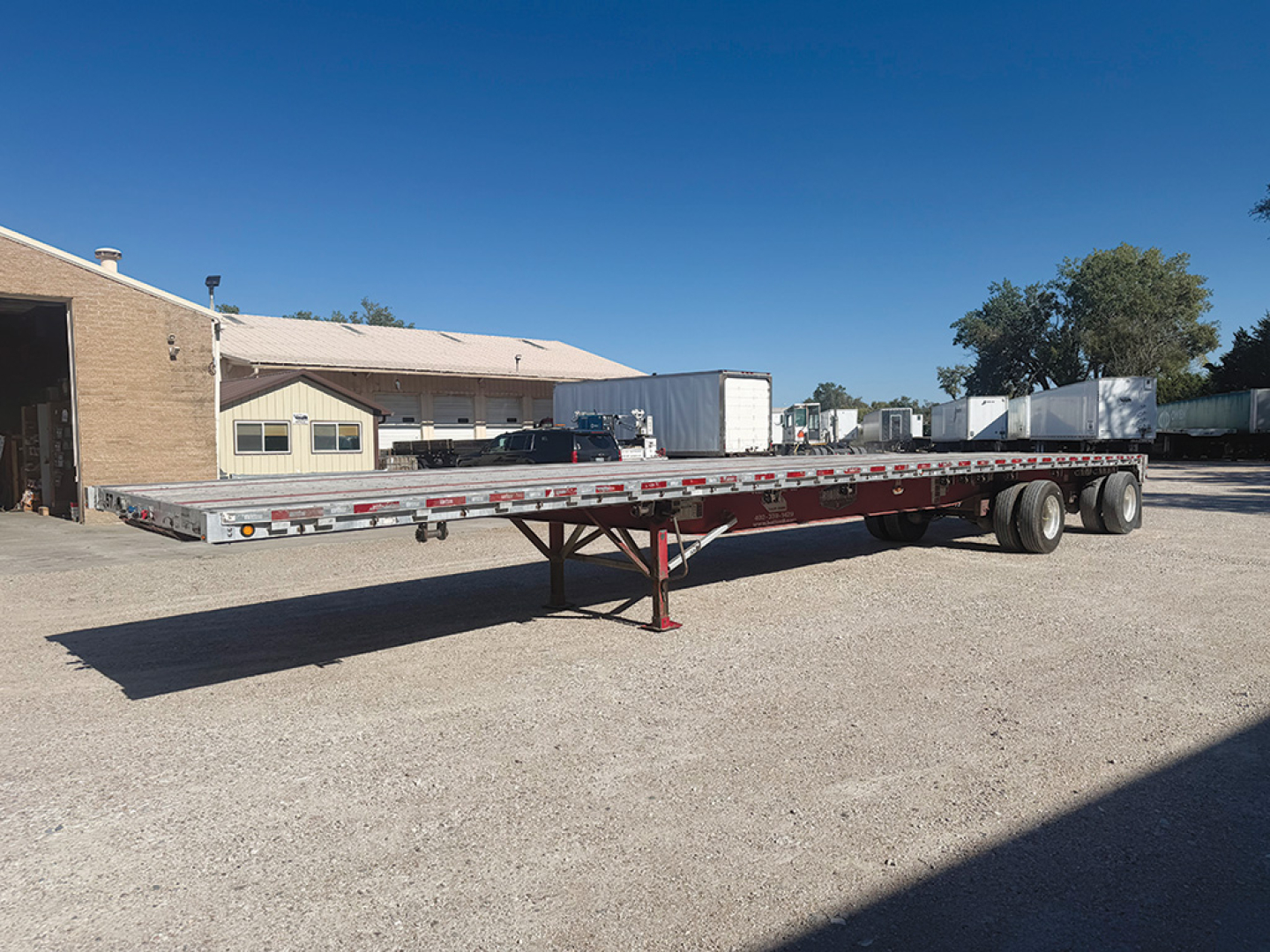 Flatbed trailer in an open lot showing a long aluminum deck with tandem axles and landing gear.