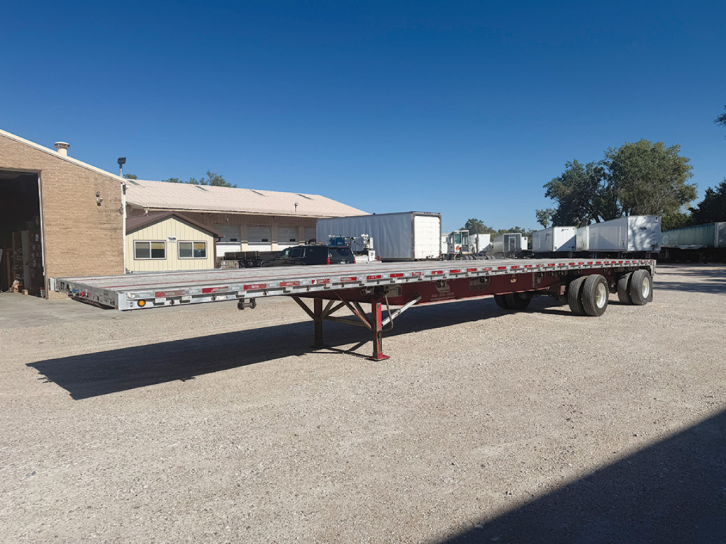 Flatbed trailer in an open lot showing a long aluminum deck with tandem axles and landing gear.