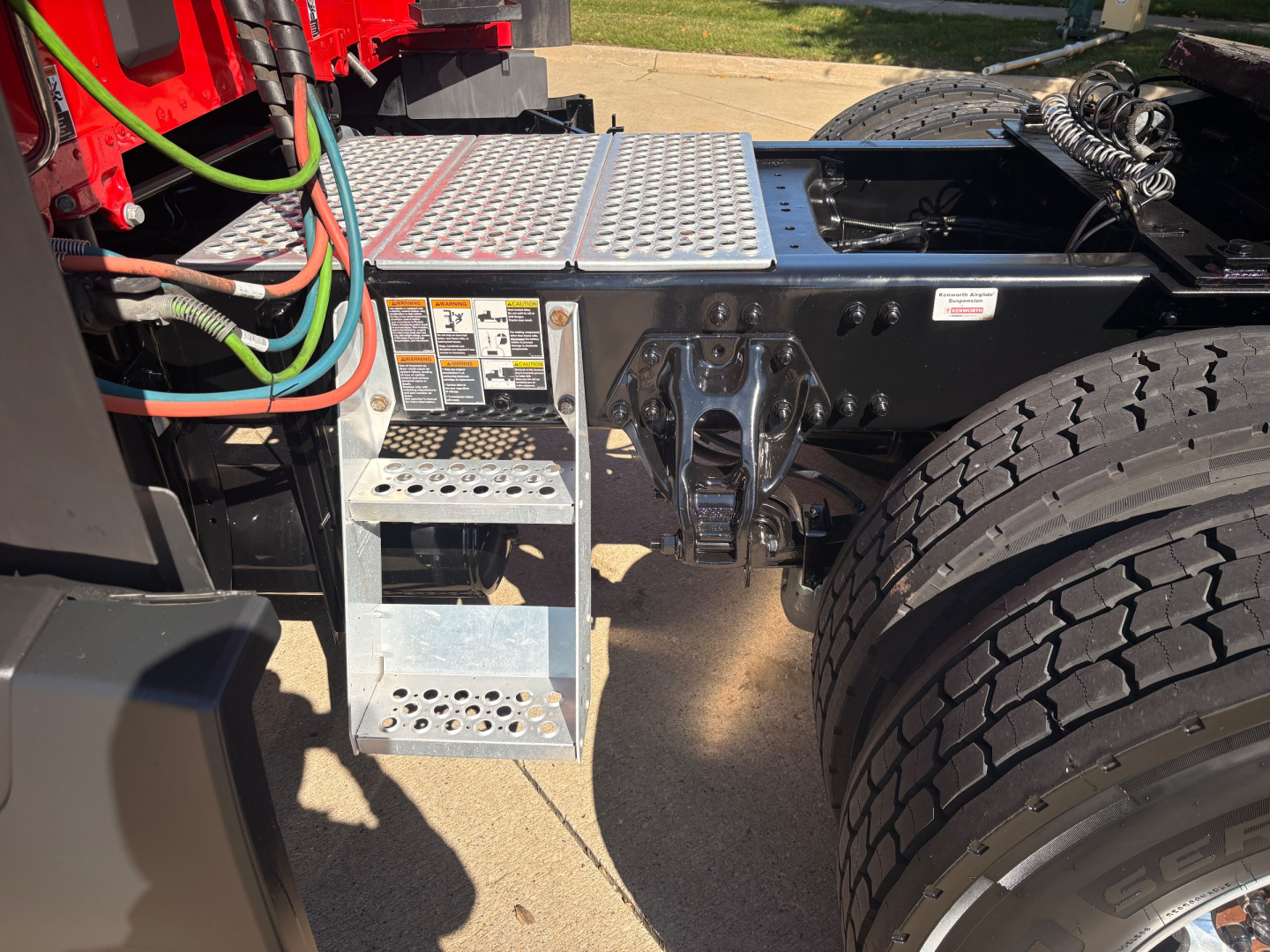 Fuel and air lines visible near the cab of a Kenworth truck. Side steps with perforated metal, battery/step area, dual rear tires, chassis rails, and hydraulic/electrical components.