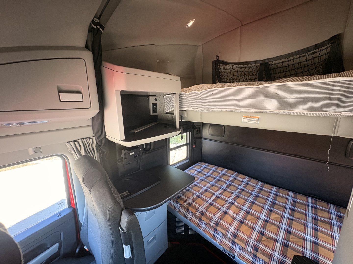 Kenworth T680 sleeper cab interior showing bunk bed with plaid sheet, overhead storage, and small desk area.