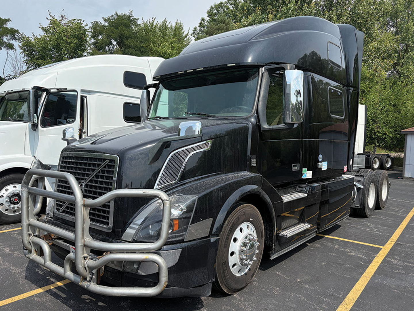 Volvo VNL 780 tractor in dark color with front bull bar, sleeper cab, large hood, chrome mirrors, and side steps; parking lot with other trucks in background.
