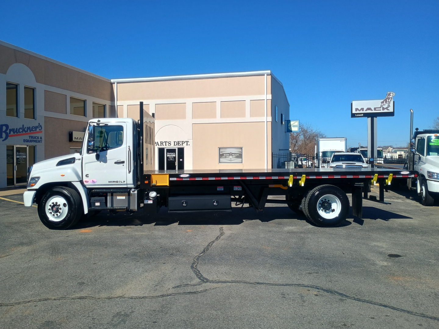 Flatbed truck side view showing 2025 Hino L7 with long flatbed in dealership lot.