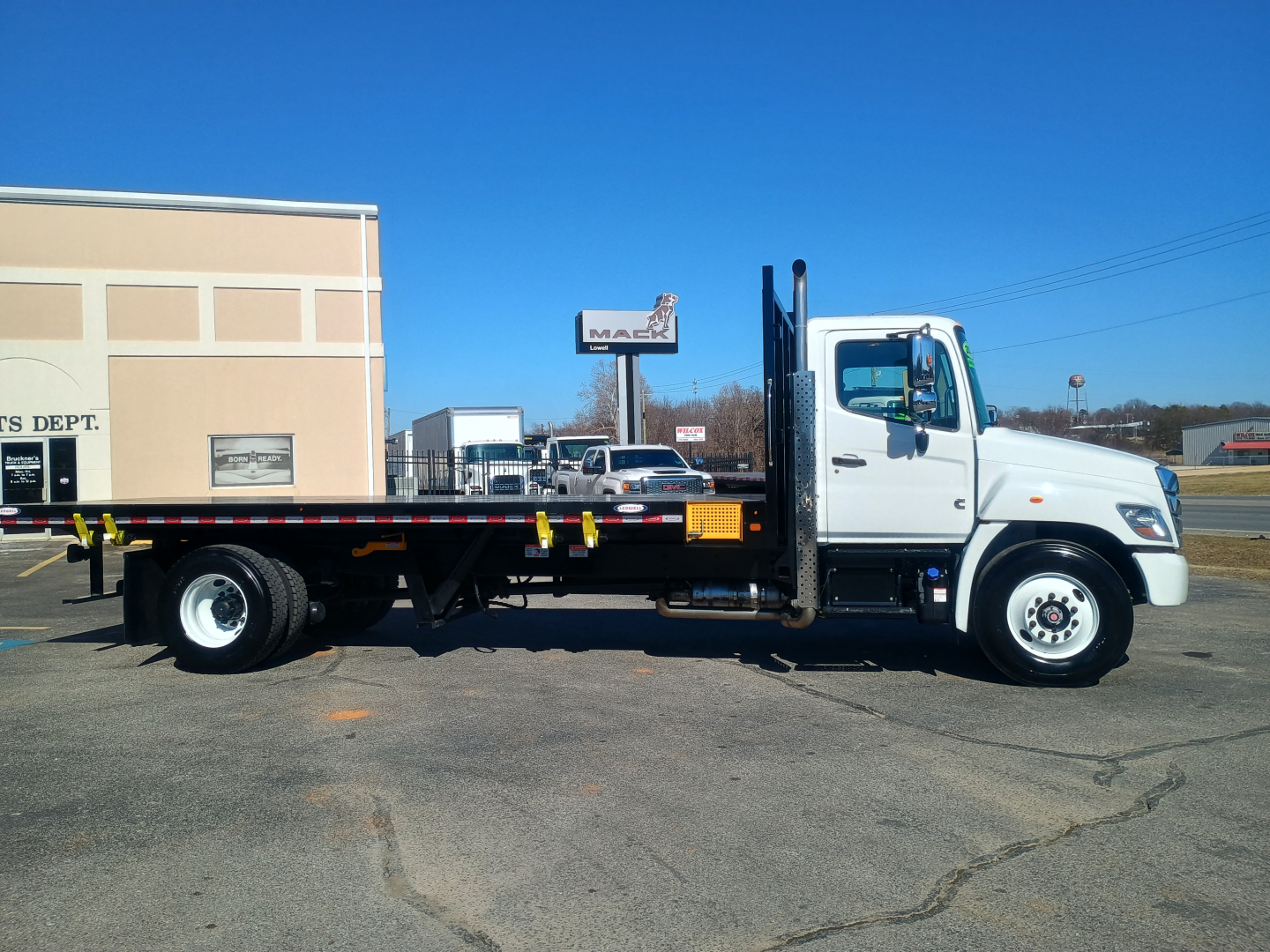 2025 Hino L7 flatbed truck side view showing cab with white paint, black flatbed, and vertical power unit ramps. Visible text includes VIN label and dealership signage in background.