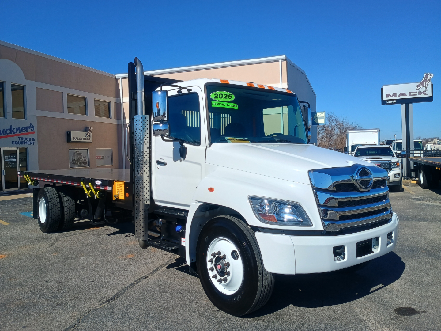 Hino L7 flatbed truck with white cab and chrome grille, stickered green 2025 on windshield, parked at dealership lot.