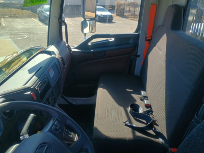 Interior cab view of a 2025 Hino L7 flatbed truck showing driver seat, dashboard, steering wheel, and co-pilot bench with seat belt buckles.