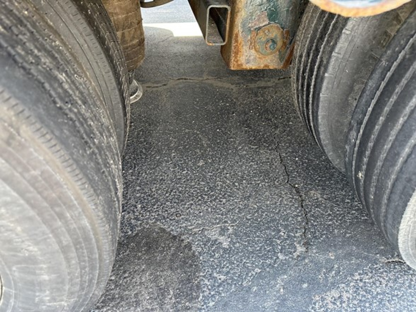 Rear view of a Trail King lowboy trailer showing dual tires and cracked asphalt surface between axles; some rusted metal hitch components visible.