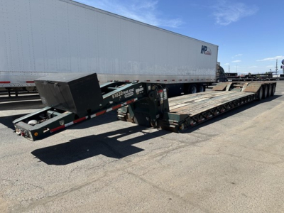 Lowboy trailer in yard with extended flatbed and gooseneck area; side rails visible; trailer and truck tires; white trailer in background; signage on gooseneck area
