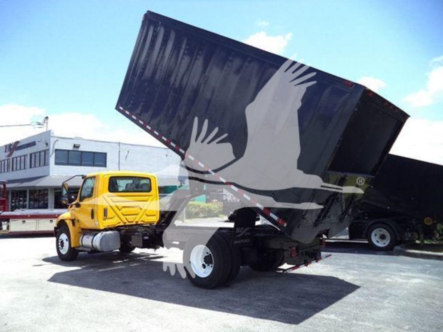 Truck with a raised dump bed, yellow cab, white wheels, and a black dumping container in an industrial lot.