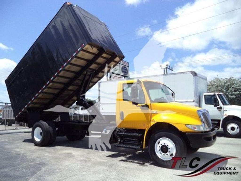 International MV truck with a raised dump bed in a yard, yellow cab, black bed tilt, front wheels, utility trucks in background.