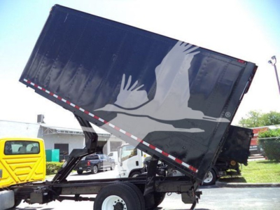 Dump truck trailer with a raised bed showing a large graphic of a white crane/dove on a dark container and a yellow truck in background.