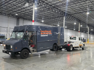 Front/side view of a dark blue delivery truck with open cab door, text on side reads 'Everything and the kitchen sink. Delivered.' The truck is in a large indoor facility with a tow vehicle attached to a white pickup tow truck.