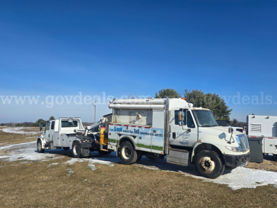 International 4300 tow truck with mounted crane/dump setup, side banner graphics, on a grassy lot with light snow.