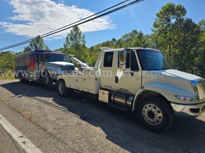 Tow truck setup showing a white International 4300 tow truck with a flatbed, connected to a larger blue tow vehicle in the background.