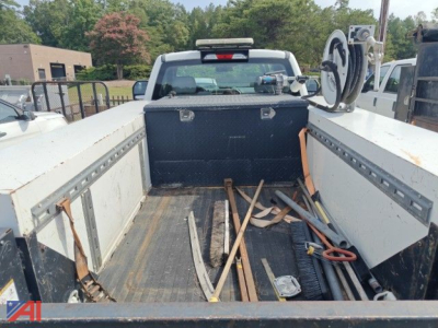 Image shows a Ford F-250 Super Duty pickup truck with a flatbed. Inside the bed are assorted metal rails, wooden boards, hoses, a black toolbox, and construction tools. Tailgate area visible, rear window, and pickup cab in background.