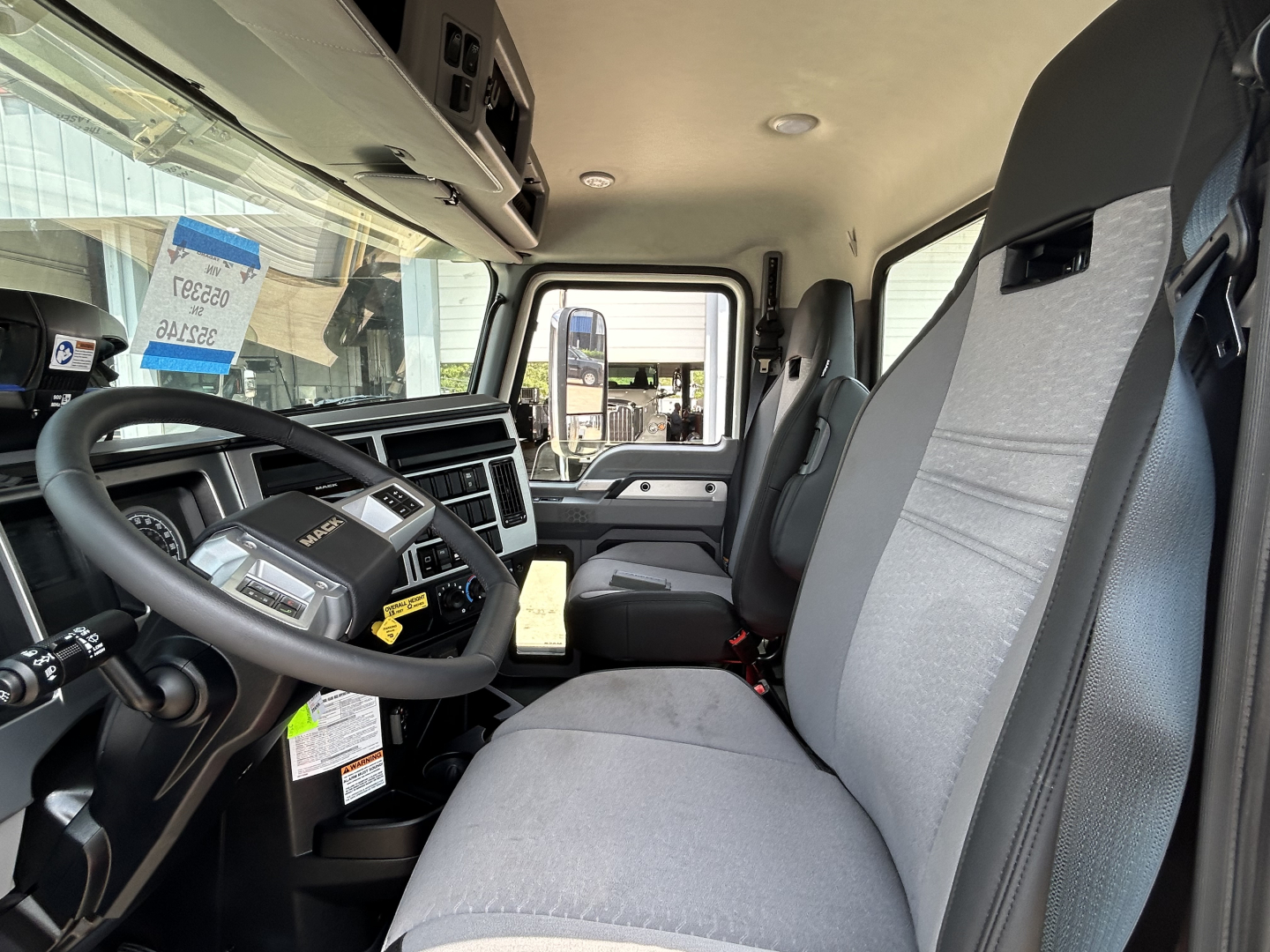 Truck cab interior of a 2027 Mack GR64F crane truck showing steering wheel, driver seat, dashboard, and passenger seat.