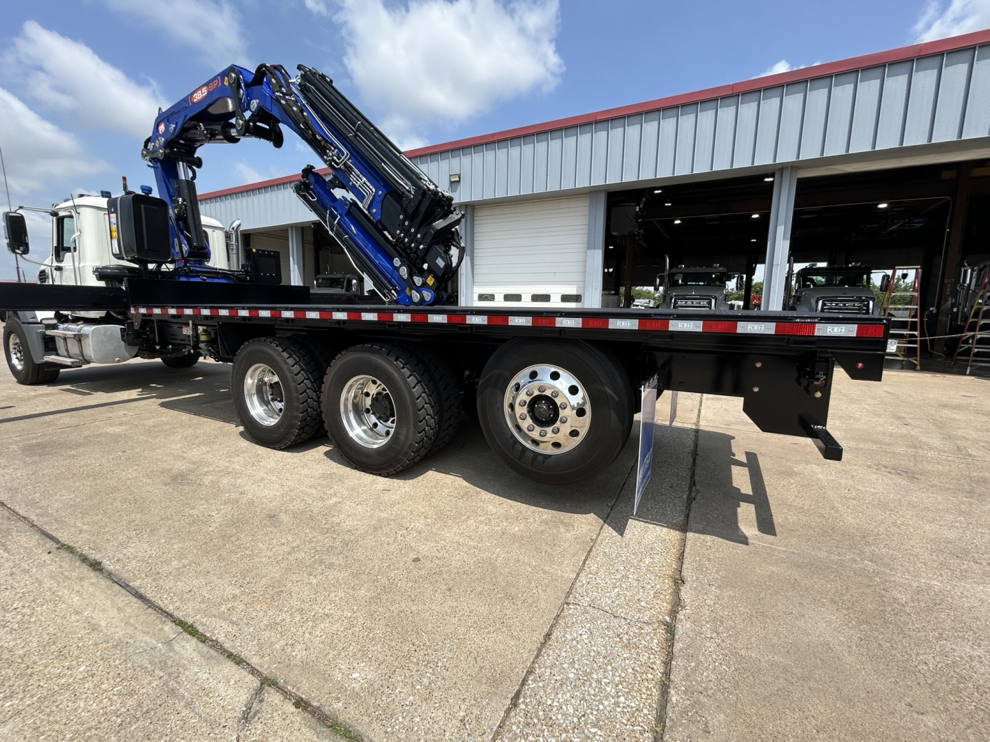Flatbed tow truck trailer with a blue crane arm, three axles, and chrome wheels. The truck is parked outside a workshop with a red roof. Visible text on the truck includes no legible labels in this view.