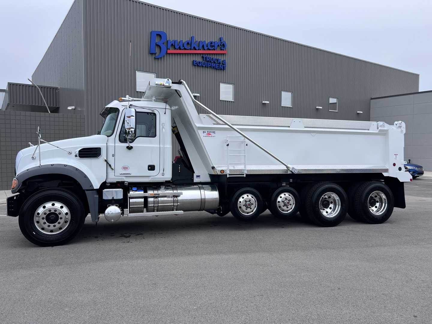 Truck side view showing 2027 Mack GR64F dump truck with white cab and dump body, hydraulic weekly hoist, six wheels, and dealer building in background