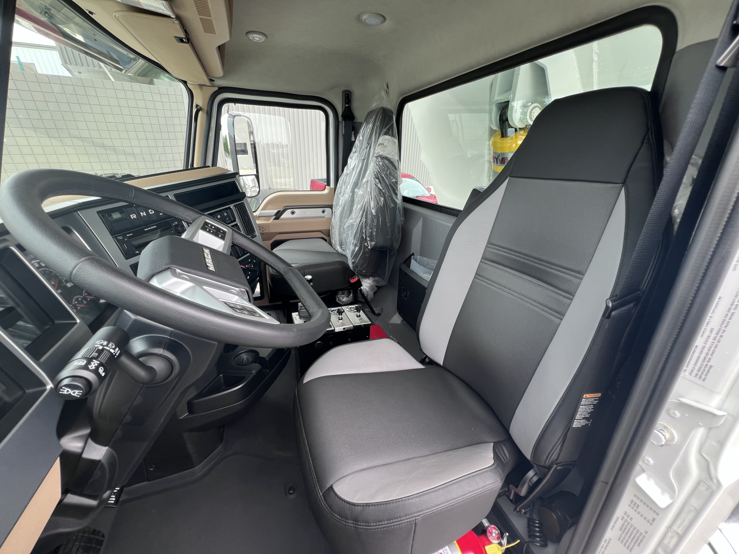 Cab interior of a 2027 Mack GR64F dump truck showing steering wheel, driver and passenger seats, and dashboard with plastic-wrapped seat cover.
