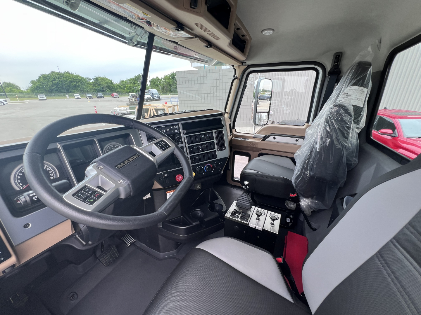 Truck cab interior showing steering wheel, dashboard with gauges, center console, and driver seat; plastic-wrapped passenger seat and gear shifter area visible.