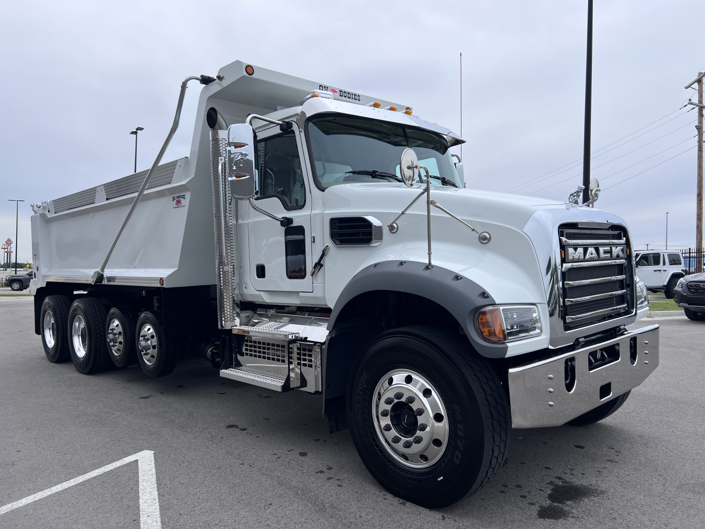 Dump truck cab and bed of a 2027 Mack GR64F showing chrome grille, dual mirrors, side steps, and multi-axle rear with dump bed