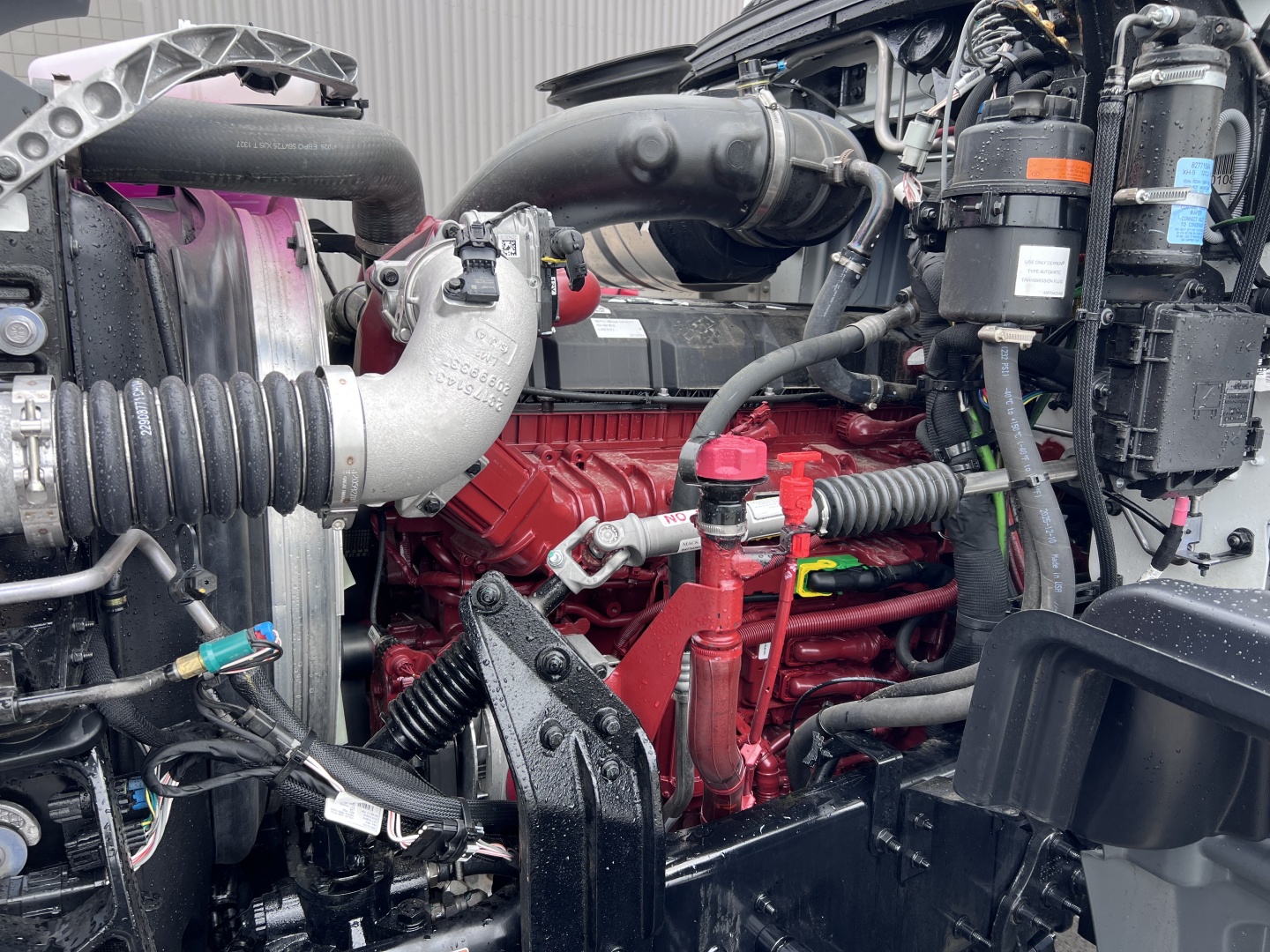 Mack GR64F engine bay showing red engine block, black hoses, air intake with corrugated hose, and surrounding components.
