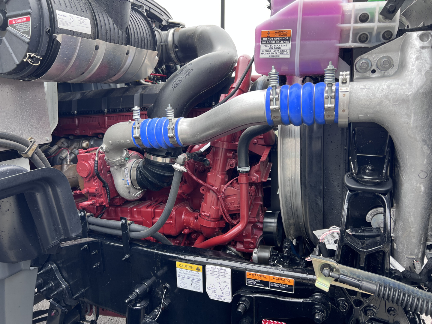 Mack GR64F truck engine bay showing red diesel engine, blue silicone hoses, black intake pipe, purple coolant reservoir, and metal radiator support.