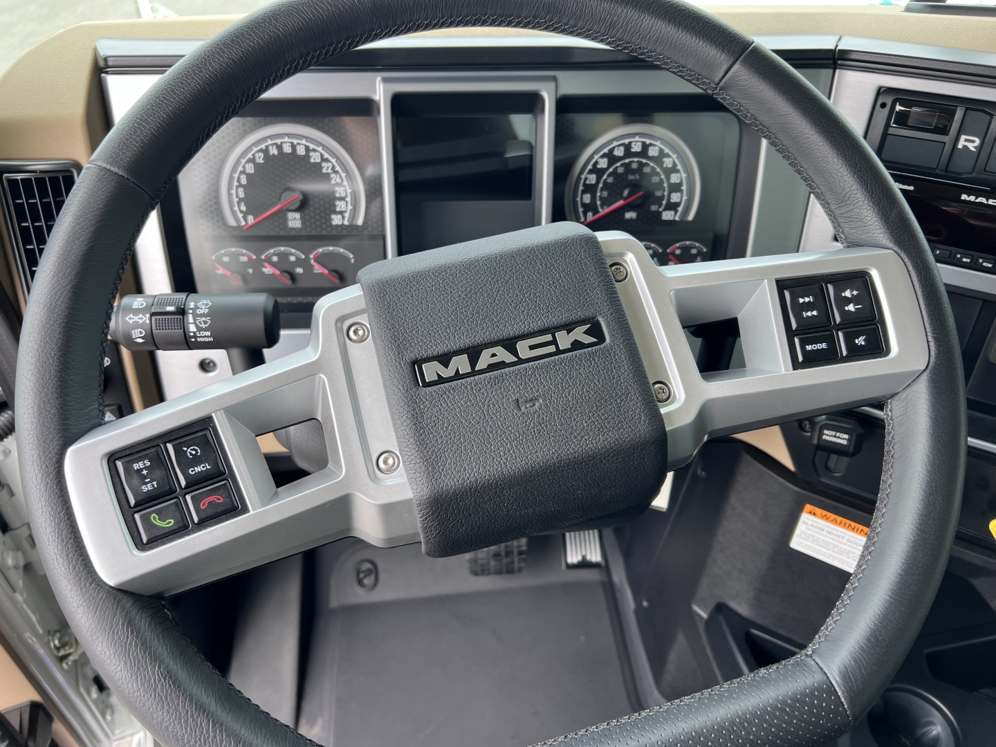 Steering wheel and dashboard of a Mack GR64F truck showing the center horn pad with MACK logo and surrounding control buttons, gauge cluster visible in background.
