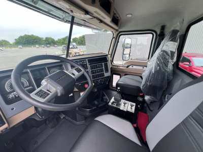 Truck cab interior showing steering wheel, dashboard with gauges, center console, and driver seat; plastic-wrapped passenger seat and gear shifter area visible.