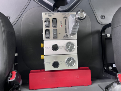 Mack GR64F cockpit controls panel with levers, switches, and dials; stacked metal panels labeled ‘PUSH TO LOAD’ and gauges, red base, part of truck interior.