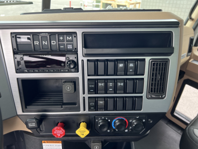 Dashboard of 2027 Mack GR64F dump truck showing gear selector panel, blank radio, assorted switches, climate controls, and push button starts.