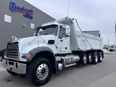 Dump truck in a Mack GR64F, front-left view, white cab, large chrome bumper, dual rear axles, dump bed with hydraulic cylinder, company building in background.