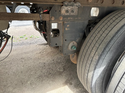 Trailer undercarriage showing frame, suspension, and dual rear axle tires with a small white label on a component and exposed wiring.