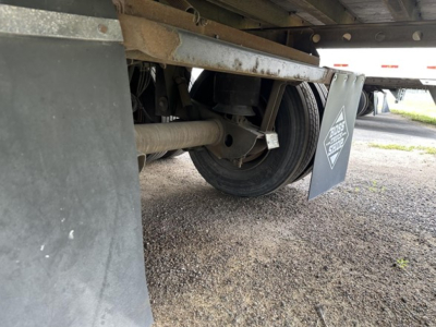 Trailer undercarriage showing wheel, suspension, and mudflap with a triangular warning sticker.
