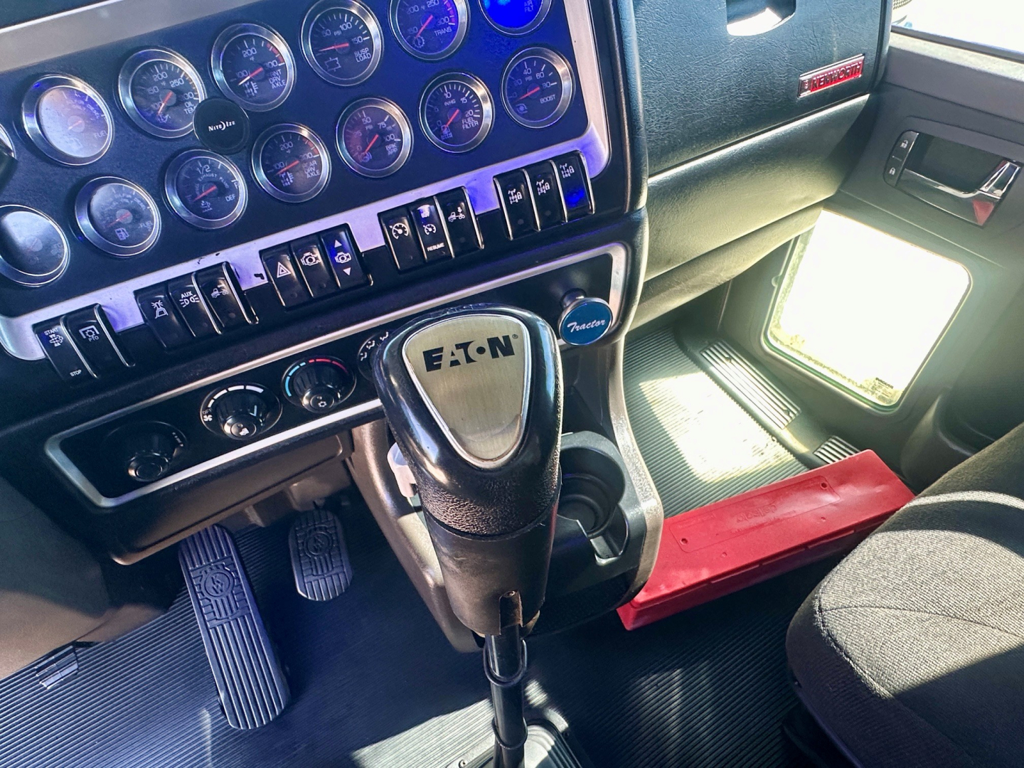 Dashboard and steering column of a Kenworth truck showing a gear selector with Eaton logo, multiple gauges, switches, and a red step under the door.