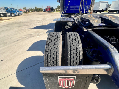 Kenworth W900 chassis rear view showing dual rear tires and frame, with logo plate on mudguard and blue cab in background.