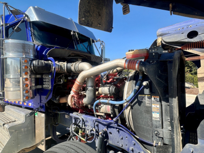 Kenworth W900 engine bay view showing turbocharger piping and various hoses and components in front of cab; blue truck with chrome exhaust, exposed engine bay.